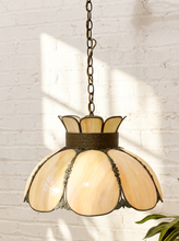 Load image into Gallery viewer, A vintage-style pendant light with a bronze metal frame and cream-colored stained glass petals, hanging against a white brick wall with soft window shadows.
