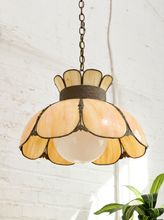 Load image into Gallery viewer, An Art Deco style pendant light with a bronze metal frame and cream-colored stained glass petals surrounding a frosted glass globe bulb, hanging against a white brick wall.
