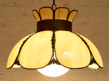 Load image into Gallery viewer, A vintage-style stained glass pendant light with a warm yellow glowing shade, featuring petal-shaped glass panels and a dark metal frame, hanging against a white brick wall.
