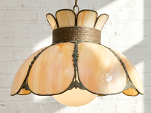 Load image into Gallery viewer, An Art Deco style pendant light with a warm peach and cream marbled glass shade, bronze metal trim, and a hanging chain, set against a white brick wall with soft sunlight and shadows.
