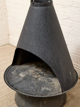 Load image into Gallery viewer, A black metal freestanding fireplace or stove with a conical hood and circular base, photographed indoors against a white brick wall and beige carpet.
