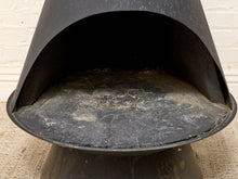 Load image into Gallery viewer, A close-up of a black metal outdoor pizza oven or stove, showing the open arched front door and a worn dark gray liner inside, set against a white brick wall and burlap floor.
