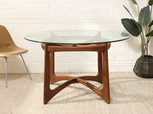 Load image into Gallery viewer, A mid-century modern round dining table with a clear glass top and a warm brown wooden base featuring sculptural curved legs, set in a room with a white brick wall, beige rug, and a potted plant.

