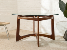 Load image into Gallery viewer, A mid-century modern wooden dining table with a clear glass top and a curved, star-shaped walnut wood base, set against a white brick wall.
