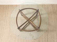 Load image into Gallery viewer, A round glass-top coffee table with a dark brown wooden geometric base featuring intersecting circular and cross-shaped elements, shown from a top-down view on a textured beige rug.
