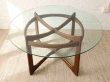 Load image into Gallery viewer, A mid-century modern round dining table with a clear glass top and a sculptural wooden base made of interlocking curved wooden arms and angled legs, set on a textured beige rug against a white brick wall.
