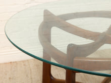 Load image into Gallery viewer, Close-up of a round glass-top table with a curved wooden frame and wooden legs, photographed indoors against a light wall and floor.
