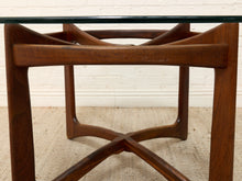 Load image into Gallery viewer, Close-up of a mid-century modern wooden table base with a dark glass top, showing the rich brown wood grain and curved geometric legs against a white brick wall and beige rug.
