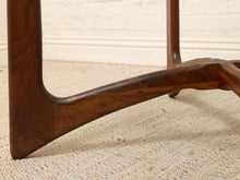 Load image into Gallery viewer, Close-up of a dark brown wooden table leg and curved support brace on a beige woven rug against a white brick wall.
