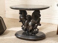 Load image into Gallery viewer, A dark carved pedestal side table featuring two lion figures supporting a round top and oval base, set on a textured rug against a white brick wall.
