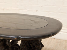 Load image into Gallery viewer, Close-up of a dark carved wooden table with a smooth oval top and ornate sculpted legs, set against a light brick wall background.
