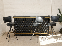 Load image into Gallery viewer, Vintage black upholstered bar with deep button tufting and brass-tone trim paired with three black swivel bar stools on metal bases
