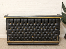 Load image into Gallery viewer, A black tufted leather bar counter with gold brass accents and a bamboo-style top and base trim, set against a white brick wall.
