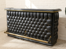 Load image into Gallery viewer, A black tufted leather bar counter with brass hardware, set against a white brick wall and beige carpet.
