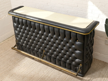 Load image into Gallery viewer, A vintage-style tufted leather bench or storage chest with a cream top, black leather upholstery, brass trim, and a brass support bar, shown in a room corner against a white brick wall.
