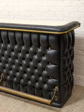 Load image into Gallery viewer, A close-up of a black tufted leather bar counter with brass hardware and a light-colored stone top, set against a white brick wall and beige carpet.
