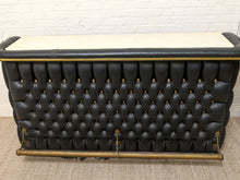 Load image into Gallery viewer, A black tufted leather bench with brass metal supports and a cream-colored top surface, set against a white brick wall and beige carpet.
