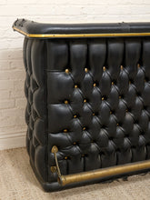 Load image into Gallery viewer, A close-up of a black tufted leather Chesterfield sofa corner with brass button tufting, a brass trim strip along the top, and a brass support rod on a beige carpet against a white brick wall.
