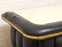 Load image into Gallery viewer, Close-up of a black leather upholstered table corner with a light wood top and brass trim, photographed against a white brick wall.
