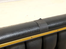 Load image into Gallery viewer, Close-up of a black textured faux-leather upholstered furniture piece with a gold metal trim strip and a black strap detail, set against a light beige background.
