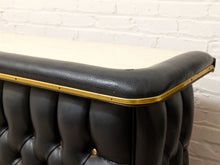 Load image into Gallery viewer, Close-up of a black tufted leather bar counter with a light wood top and brass trim, set against a white brick wall.
