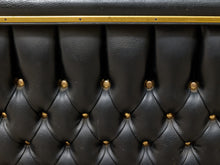 Load image into Gallery viewer, Close-up of a black tufted leather headboard with gold brass buttons and a gold horizontal trim strip.
