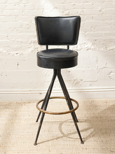 Load image into Gallery viewer, A black faux-leather swivel barstool with a rectangular backrest, distressed black metal legs, and a circular bronze footrest ring, set against a white brick wall and beige carpet.
