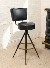 Load image into Gallery viewer, A vintage-style bar stool with a black leather upholstered seat and backrest, supported by a distressed black metal tripod base with a brass foot ring, set on a beige carpet against a white brick wall.
