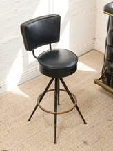 Load image into Gallery viewer, A vintage-style bar stool with a black faux-leather swivel seat and backrest, supported by a distressed black metal frame with a circular footrest ring, set on a beige textured rug against a white brick wall.
