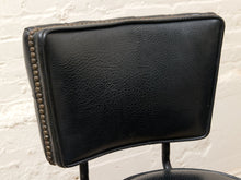 Load image into Gallery viewer, Close-up of a black leather chair backrest with a pebbled texture and nailhead trim, set against a white brick wall.
