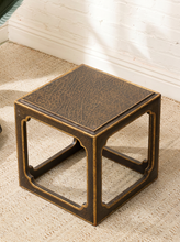 Load image into Gallery viewer, A small cube-shaped side table with a distressed bronze finish and a textured leather-look top, shown on a beige carpeted floor against a white brick wall.
