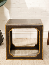 Load image into Gallery viewer, A square dark brown wooden side table with a distressed finish and gold-painted trim, featuring a large open center panel with decorative curved corners, set on a beige woven rug against a white brick wall.
