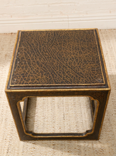 Load image into Gallery viewer, A square decorative side table or stool with a dark brown and black crackled enamel top and gold-toned metal frame, set on a woven rug against a white brick wall.
