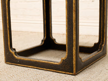 Load image into Gallery viewer, Close-up of a dark brown wooden side table or stool base with a distressed finish and gold leaf accents, set on a textured beige rug against a white brick wall.
