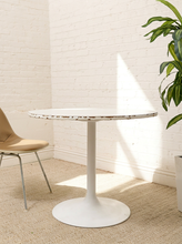 Load image into Gallery viewer, A white round pedestal dining table with a distressed, chipped-edge top surface, set in a bright room with a white brick wall, beige rug, a modern chair, and a green plant.
