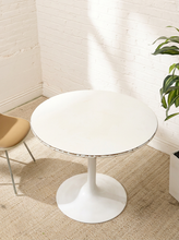 Load image into Gallery viewer, A round white pedestal dining table with a tulip base, set on a textured beige rug in a bright room with a white brick wall, a partial tan chair on the left, and a green plant on the right.
