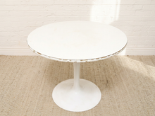 Load image into Gallery viewer, A round white dining table with a tulip base, set on a textured beige rug against a white brick wall.
