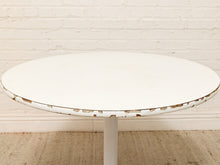 Load image into Gallery viewer, A round white table with a distressed finish showing brown wood underneath the chipped paint, supported by a white pedestal base against a white brick wall and beige rug.
