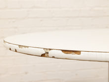 Load image into Gallery viewer, Close-up of a round white table with heavily distressed and peeling paint along the rim, set against a white painted brick wall background.
