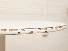 Load image into Gallery viewer, A close-up of a distressed white round table showing chipped and peeling paint along the rim, set against a white painted brick wall background.
