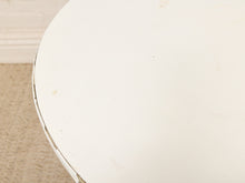 Load image into Gallery viewer, A close-up detail shot of a round white table top with a distressed finish, showing minor scuffs and scratches, with a beige carpet and white baseboard visible in the background.
