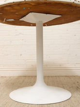 Load image into Gallery viewer, A white tulip pedestal table base with a round wooden top, shown against a white brick wall and beige rug.
