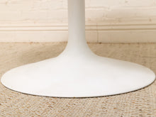 Load image into Gallery viewer, Close-up of a white tulip-style table base on a woven beige rug against a light brick wall.
