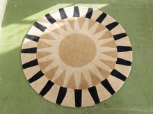 Load image into Gallery viewer, A round area rug with a sunburst pattern, featuring a beige center, tan rays, and alternating black and beige stripes in the outer border, laid flat on a green floor.
