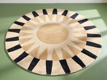 Load image into Gallery viewer, A round area rug with a sunburst pattern in beige and black, displayed on a solid sage green floor.
