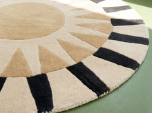 Load image into Gallery viewer, A close-up of a round area rug with a sunburst design, featuring a beige center, tan rays, and alternating cream and black stripes along the outer rim, shown on a green surface.

