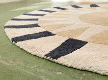 Load image into Gallery viewer, A close-up of a round area rug with a sunburst pattern in cream, tan, and dark charcoal, shown on a distressed green floor.
