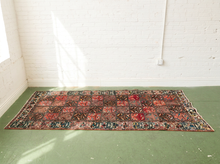 Load image into Gallery viewer, A rectangular vintage-style area rug with a complex geometric and floral pattern in red, blue, black, and beige, laid out on a green floor against a white brick wall.
