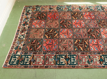 Load image into Gallery viewer, A square vintage Persian-style area rug with a geometric patchwork design in red, brown, beige, and blue, laid out on a green floor.
