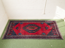 Load image into Gallery viewer, A rectangular vintage-style area rug with a red central field, dark navy and black borders, and small floral motifs, displayed on a green floor against a white brick wall.
